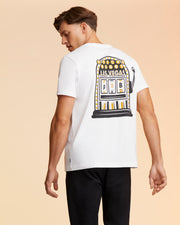 A man with brown hair is facing away from the camera, wearing a white MENS VEGAS CITY TEE - B6U967X1PC by Psycho Bunny with a colorful graphic of a Las Vegas-themed slot machine on the back. The background is a gradient of peach to beige tones. He is also wearing black pants.