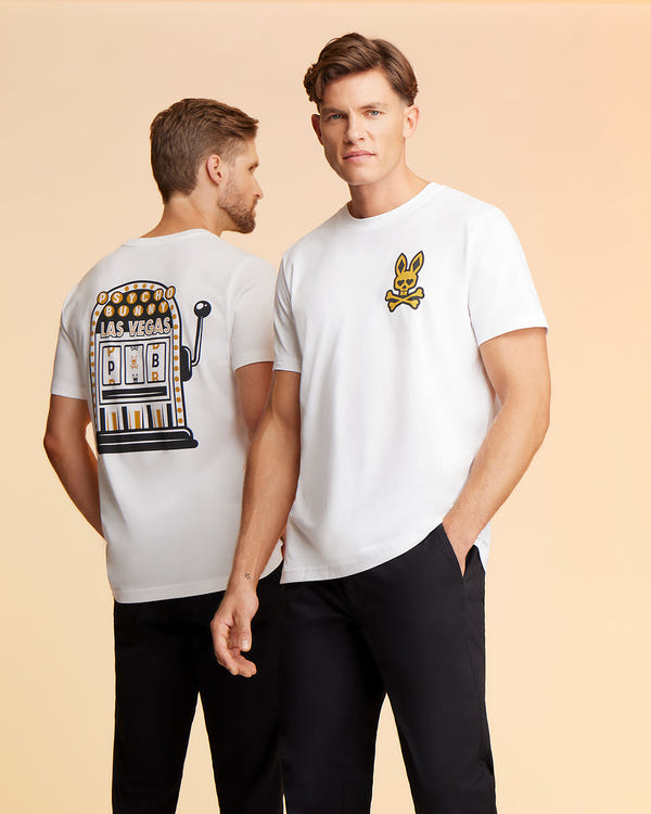 Two men wear white t-shirts and black pants. The man on the left faces away, showcasing a Psycho Bunny men's Vegas City tee - B6U967X1PC with a slot machine graphic. The man on the right, in a Pima cotton jersey, faces forward, his t-shirt featuring a small Printed Bunny Logo on the chest.