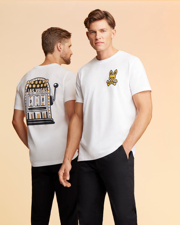 Two men wear white t-shirts and black pants. The man on the left faces away, showcasing a Psycho Bunny men's Vegas City tee - B6U967X1PC with a slot machine graphic. The man on the right, in a Pima cotton jersey, faces forward, his t-shirt featuring a small Printed Bunny Logo on the chest.