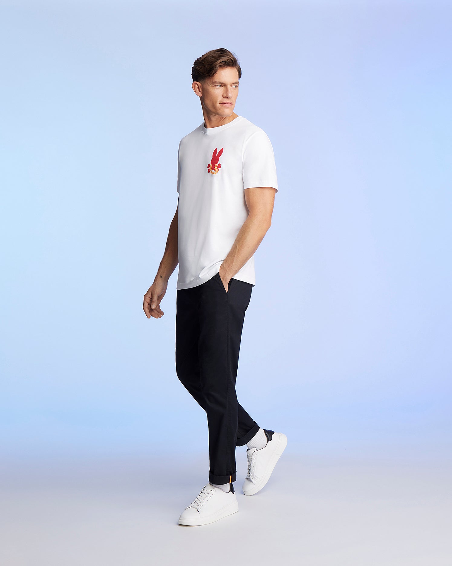 A man in a Psycho Bunny MENS HAWAII CITY TEE - B6U169Y1PC with a red graphic pattern on the chest and black pants stands against a plain, light blue background. He is looking to his left and walking while his right hand is relaxed by his side. He is wearing white sneakers.