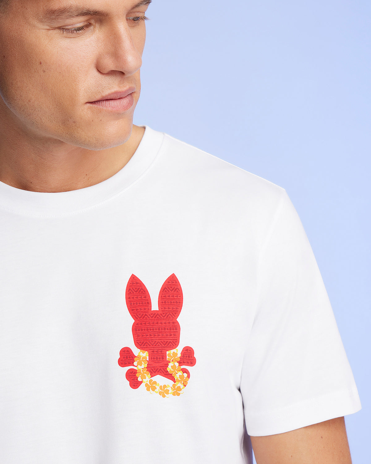 A person wearing a white Psycho Bunny MENS HAWAII CITY TEE - B6U169Y1PC with a red, bunny-shaped design adorned with yellow and orange flowers on the chest. The background is light blue, and the person's face is partially visible, looking down and to the side.