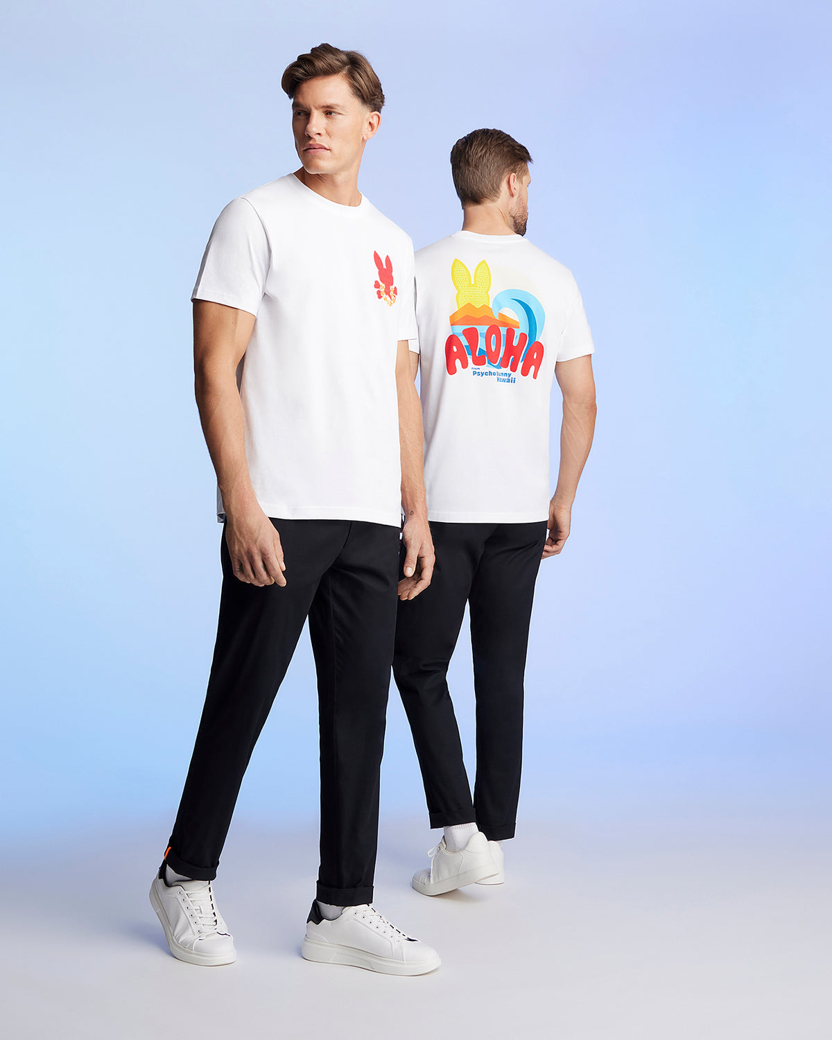 Two men are standing against a light blue background, wearing matching white t-shirts and black pants. The man on the left faces the camera, showcasing a red graphic pattern on his Pima cotton jersey. The man on the right is turned away, revealing a colorful "ALOHA" Hawaii tee design on the back of his MENS HAWAII CITY TEE - B6U169Y1PC by Psycho Bunny.