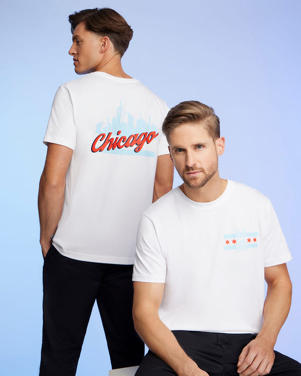 Two men are wearing Psycho Bunny's MENS CHICAGO CITY TEE - B6U551W1PC in white Pima cotton jersey. The man on the left showcases the back of his tee, which features a "Chicago" design along with a skyline graphic, while the man on the right is seated, highlighting a front graphic that includes a city-inspired bunny logo. Both tees are styled with black pants.
