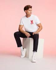 A man sits on a white rectangular block against a pink background. He is wearing a Psycho Bunny MENS NEW YORK TEE - B6U554W1PC made from Pima cotton jersey featuring a red bunny and crossbones graphic, black pants, white socks, and white sneakers. His hair is styled neatly, and he looks off to the side.