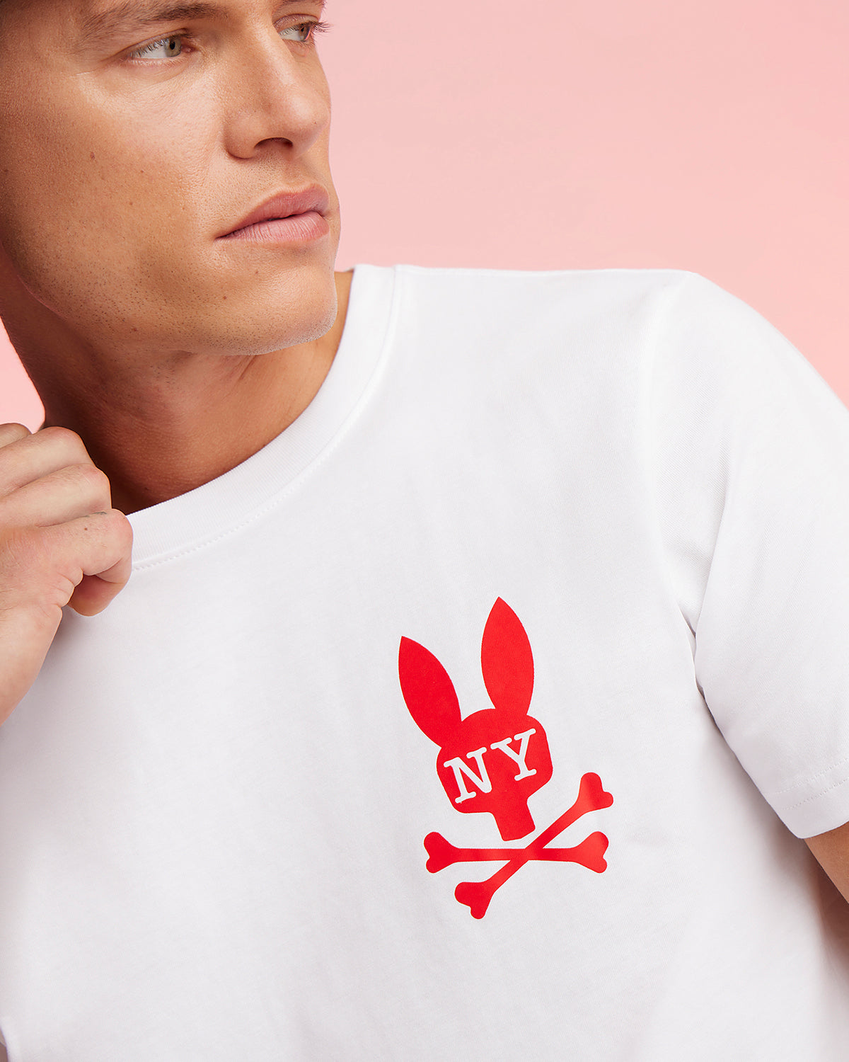 A person wearing a Psycho Bunny MENS NEW YORK TEE - B6U554W1PC made of soft Pima cotton jersey, featuring a red graphic bunny logo above crossed bones and "NY" on the bunny's head. They're looking to the side against a light pink background.