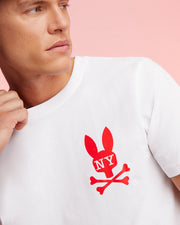 A person wearing a Psycho Bunny MENS NEW YORK TEE - B6U554W1PC made of soft Pima cotton jersey, featuring a red graphic bunny logo above crossed bones and "NY" on the bunny's head. They're looking to the side against a light pink background.