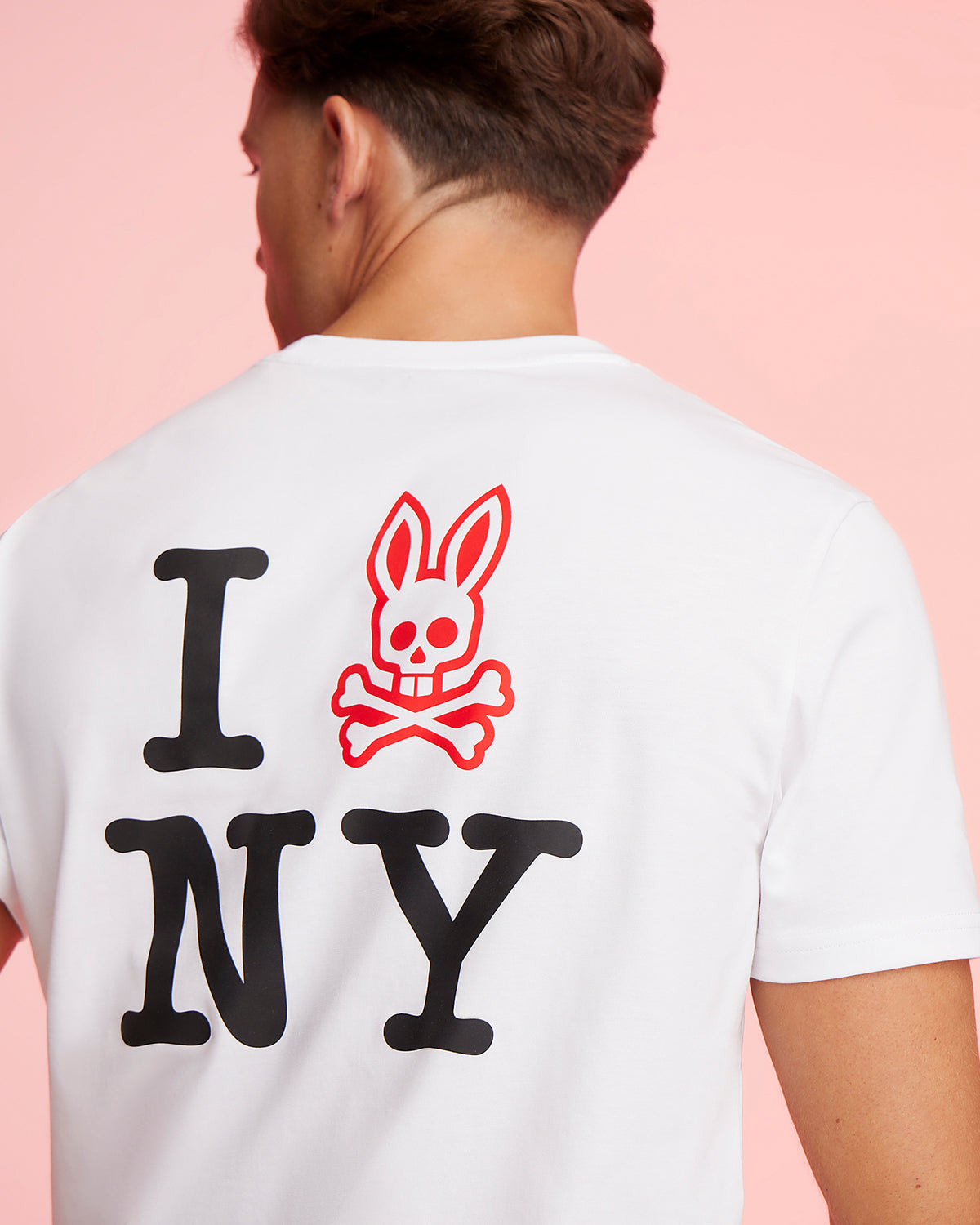 A person is shown from the back wearing a white Psycho Bunny MENS NEW YORK TEE - B6U554W1PC adorned with an "I 🐰 NY" design. Instead of a heart, there's a red bunny head with crossbones graphic. The background is a solid light pink color.