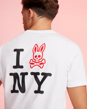 A person is shown from the back wearing a white Psycho Bunny MENS NEW YORK TEE - B6U554W1PC adorned with an "I 🐰 NY" design. Instead of a heart, there's a red bunny head with crossbones graphic. The background is a solid light pink color.