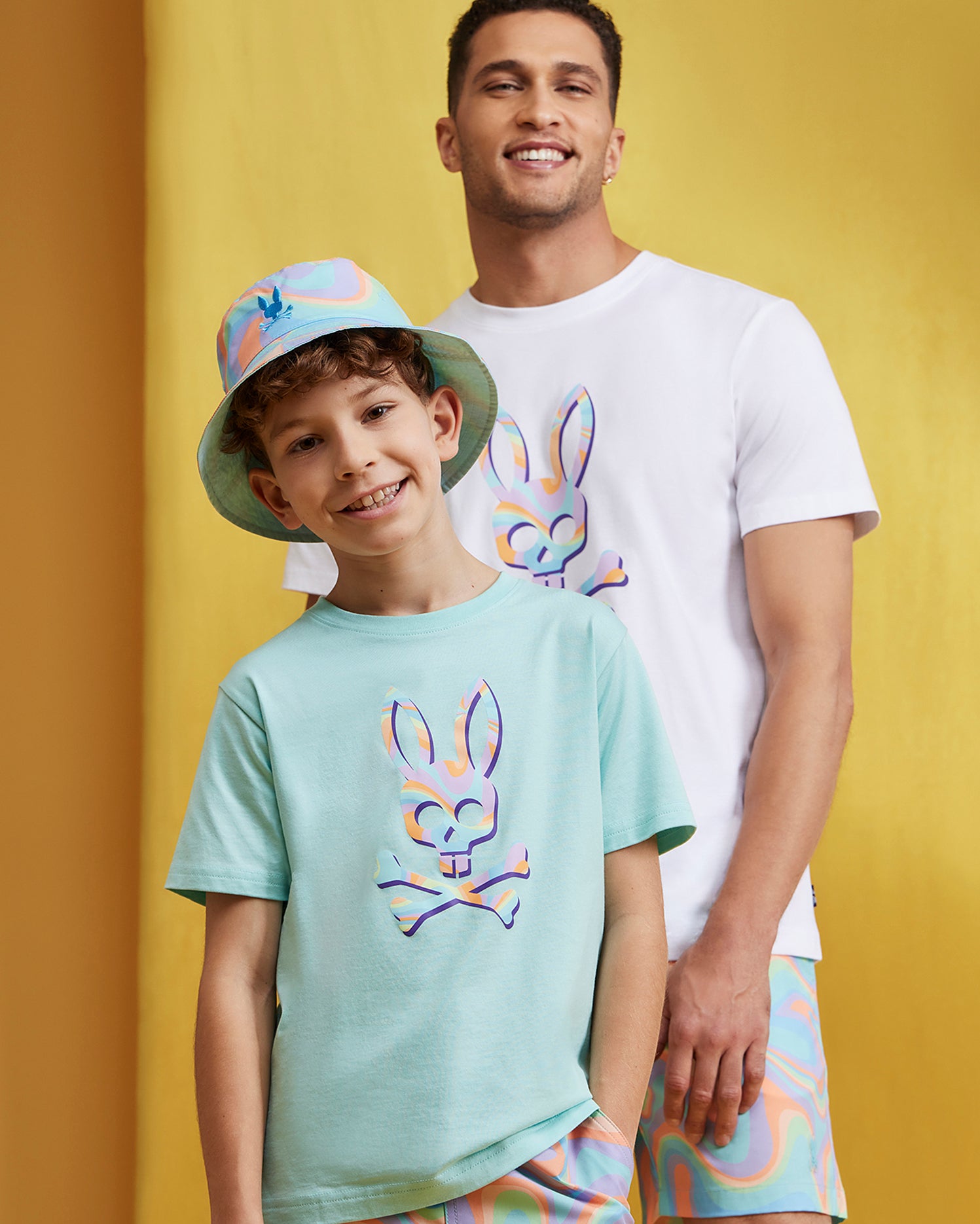 A smiling boy wearing a mint green t-shirt and a multicolored bucket hat stands in front of a man sporting a white t-shirt. Both shirts feature a colorful bunny skull graphic, with the boy pairing his look with quick-drying Psycho Bunny KIDS BLOOMINGTON SWIM TRUNK - B0W941A2PO. The background is a solid yellow.