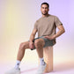 A man sits casually on a wooden stool, wearing a Psycho Bunny Men's Classic Crew Neck Tee in beige, gray shorts, and white sneakers, with a soft colorful gradient background.