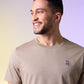 A man smiling and looking to the side, wearing a beige Psycho Bunny men's classic crew neck tee with a small Psycho Bunny embroidered logo on the chest, set against a soft gradient background.