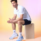 A man in sportswear, including a white Psycho Bunny Pima cotton polo and blue shorts, squats on a wooden box against a soft pink and yellow gradient background. He looks to the side thoughtfully.