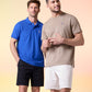 Two men standing side by side, one in a blue polo shirt with a Psycho Bunny embroidered logo and black shorts, the other in a beige t-shirt and khaki shorts, against a soft colored background.