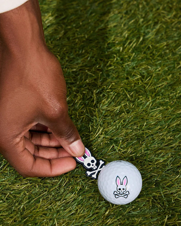 A hand places a Psycho Bunny BALL MARKER - B6A698C200, adorned with a white bunny skull and crossbones, on grass next to a matching bunny skull and crossbones golf tee from the golfers collection.