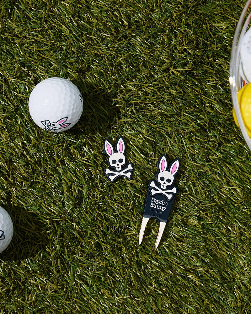 Golf balls and a divot tool from the Psycho Bunny golfer's collection, featuring the iconic skull and crossbones with bunny ears, are placed on a grassy lawn. A bowl partially visible on the right contains yellow golf balls, while a white BALL MARKER - B6A698C200 with a bunny face rests nearby.