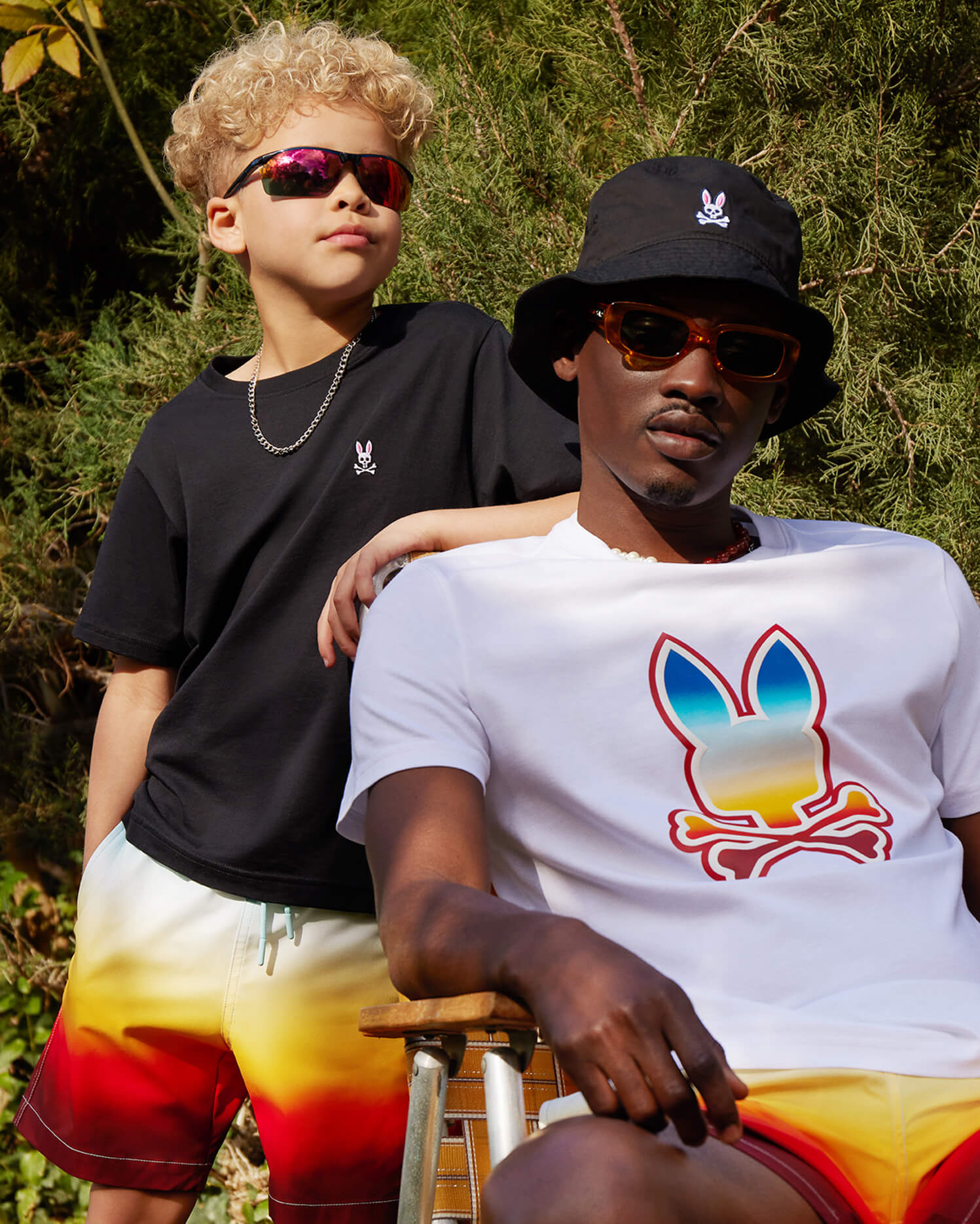 A child wearing reflective sunglasses and vibrant sunrise-inspired KIDS GUY SWIM TRUNK by Psycho Bunny is alongside an adult in colorful summer outfits, complete with sunrise prints on tees and shorts. The adult sports a black bucket hat and sunglasses, set against a backdrop of trees outdoors.