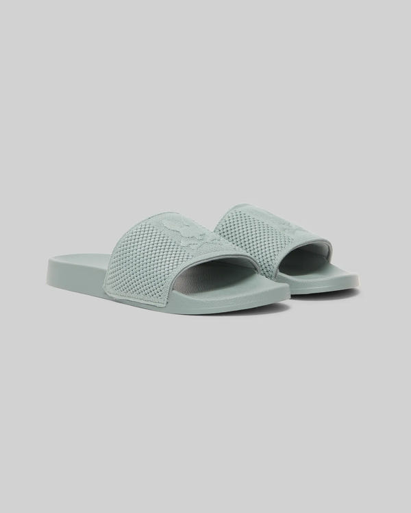 The Psycho Bunny KIDS KNIT SLIDES - B0A377F200 are light blue-green knit textile pool slides with textured straps and an ergonomically molded footbed, displayed on a plain light gray background.