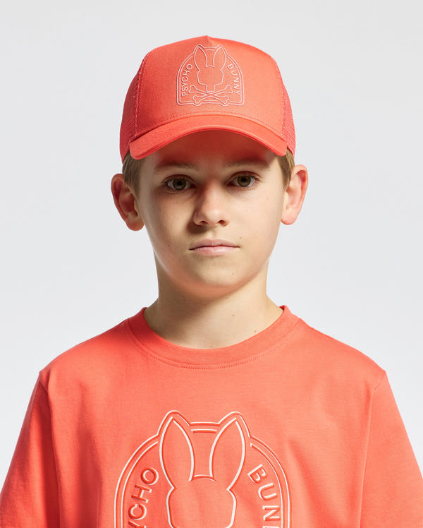 A young boy wears a matching coral Psycho Bunny T-shirt and KIDS MELBOURNE GRAPHIC TRUCKER HAT, both with an embroidered rabbit icon. The hat's snap-back strap adds style to the plain white background.