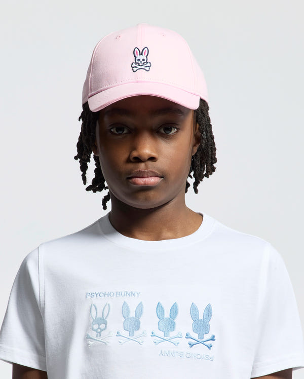 A young kid wears the pink Psycho Bunny Kids Classic Baseball Cap - B0A816A1HT, pairing it with a white T-shirt adorned with bunny designs and "Psycho Bunny" text. The adjustable cap offers comfort, while the light background enhances their playful style.