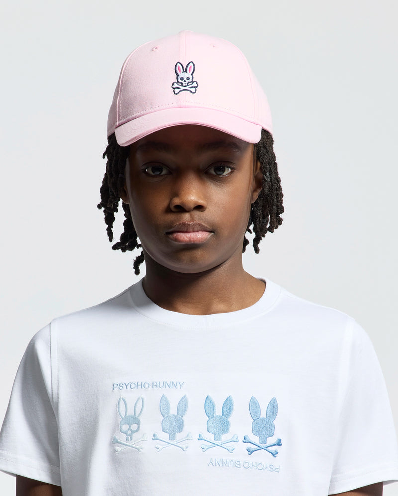 A young kid wears the pink Psycho Bunny Kids Classic Baseball Cap - B0A816A1HT, pairing it with a white T-shirt adorned with bunny designs and "Psycho Bunny" text. The adjustable cap offers comfort, while the light background enhances their playful style.