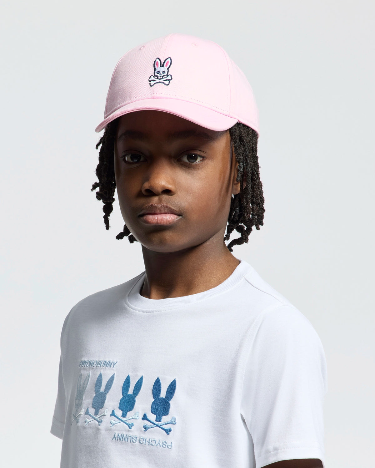 A person in a Psycho Bunny KIDS CLASSIC BASEBALL CAP - B0A816A1HT, showcasing an embroidered bunny logo, and a white T-shirt with multiple bunny designs. With braided hair and a neutral expression, they stand against a plain backdrop.