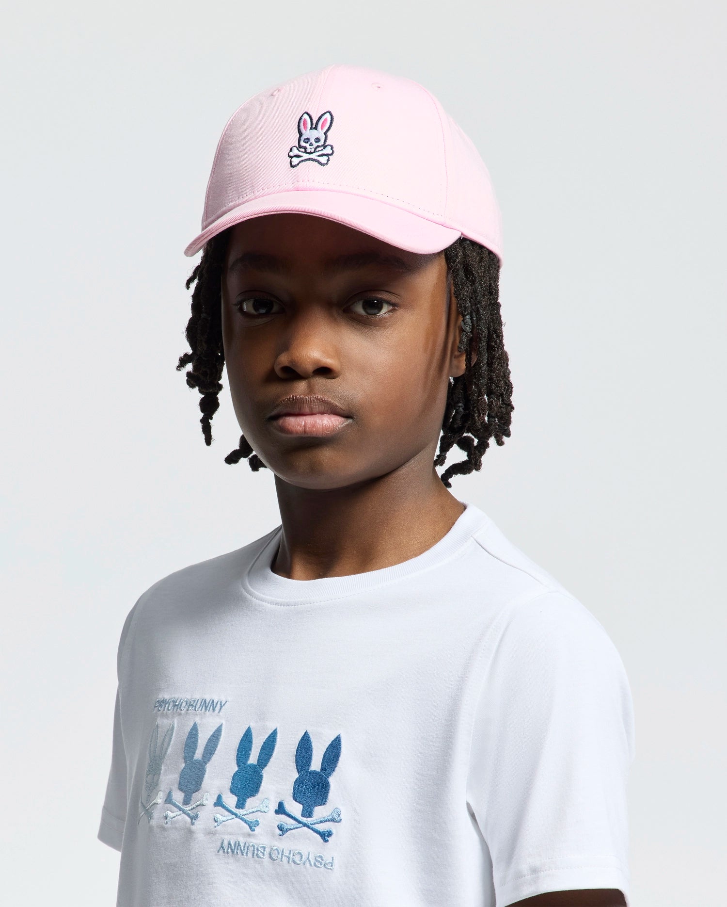 A person in a Psycho Bunny KIDS CLASSIC BASEBALL CAP - B0A816A1HT, showcasing an embroidered bunny logo, and a white T-shirt with multiple bunny designs. With braided hair and a neutral expression, they stand against a plain backdrop.