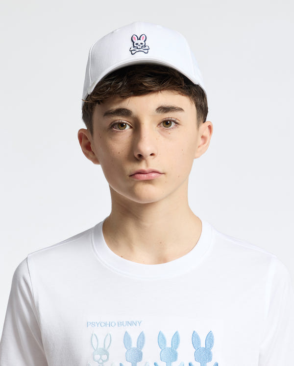 A young person is wearing a Psycho Bunny KIDS CLASSIC BASEBALL CAP in white featuring an embroidered bunny and crossbones logo, with an adjustable fit, paired with a white shirt covered in multiple bunny designs against a plain white background.