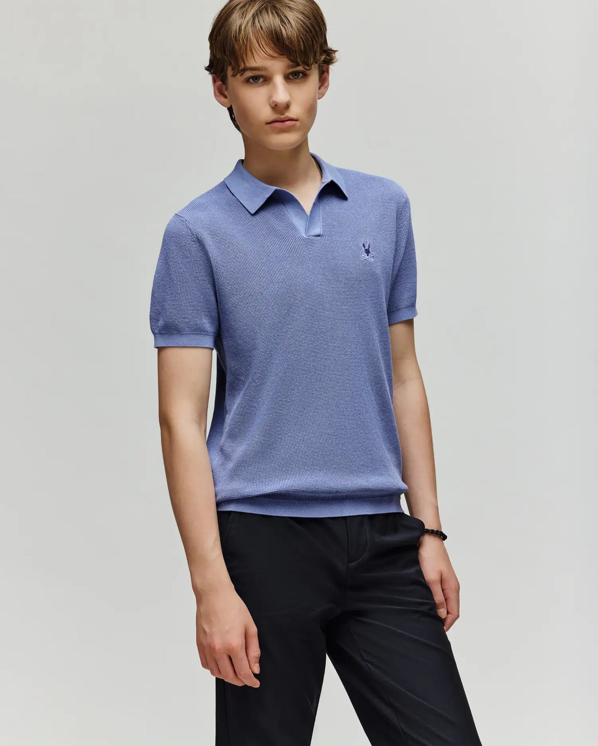 A young person with short brown hair wears a light blue Psycho Bunny KIDS LEETON KNIT POLO SHIRT (B0E562F200) with black pants, standing against a plain light background and showing a neutral expression.