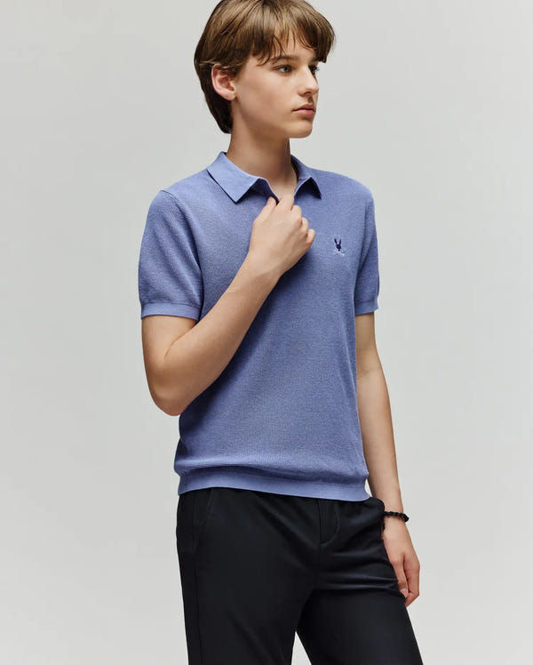 A young person with short brown hair stands against a light background, wearing the Psycho Bunny KIDS LEETON KNIT POLO SHIRT (B0E562F200) in light blue with a Johnny collar and dark pants, one hand touching their collar.