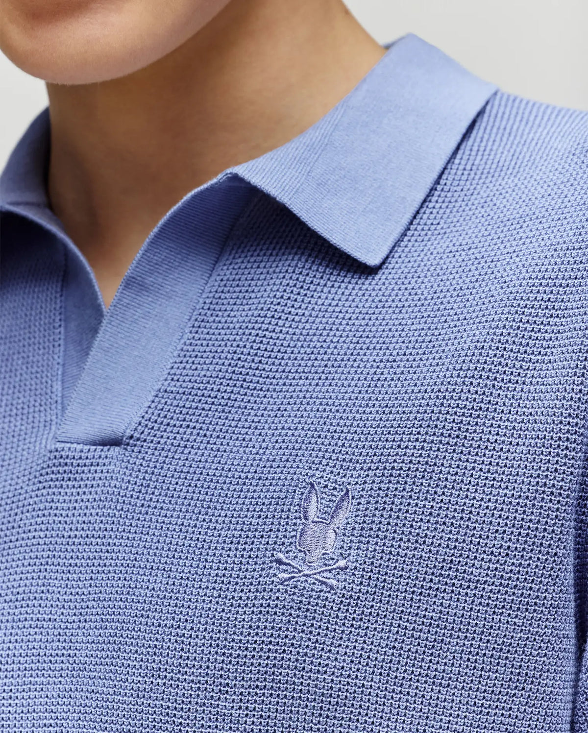 Close-up of a child wearing the Psycho Bunny KIDS LEETON KNIT POLO SHIRT (B0E562F200), featuring textured light blue cotton, a Johnny collar, and an embroidered bunny logo on the chest. Only the shirt, neck, and chin are visible.