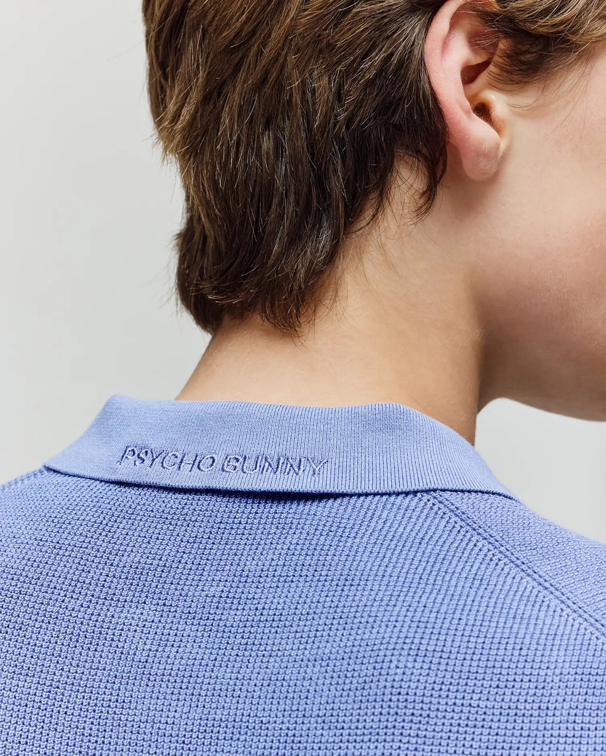 Close-up of the back of a child’s neck and head with short brown hair, showing the collar of a textured light blue Psycho Bunny KIDS LEETON KNIT POLO SHIRT (B0E562F200), embroidered with “Psycho Bunny.”.