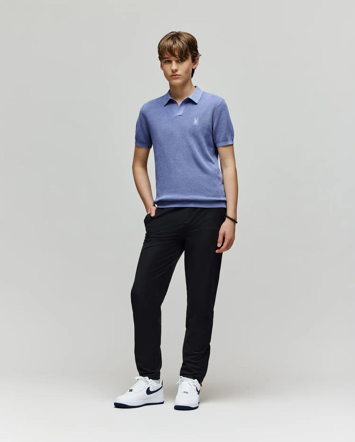 A young person with short light brown hair and a neutral expression stands against a light gray background, wearing a Psycho Bunny KIDS LEETON KNIT POLO SHIRT - B0E562F200 in light blue, black pants, and white Nike sneakers.