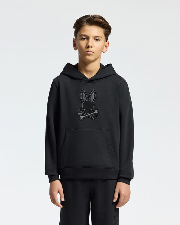 A young person stands in a black Psycho Bunny KIDS NEW CASTLE FRENCH TERRY HOODIE, style B0H517F200, featuring an embroidered bunny and crossbones design on the front against a plain white background.