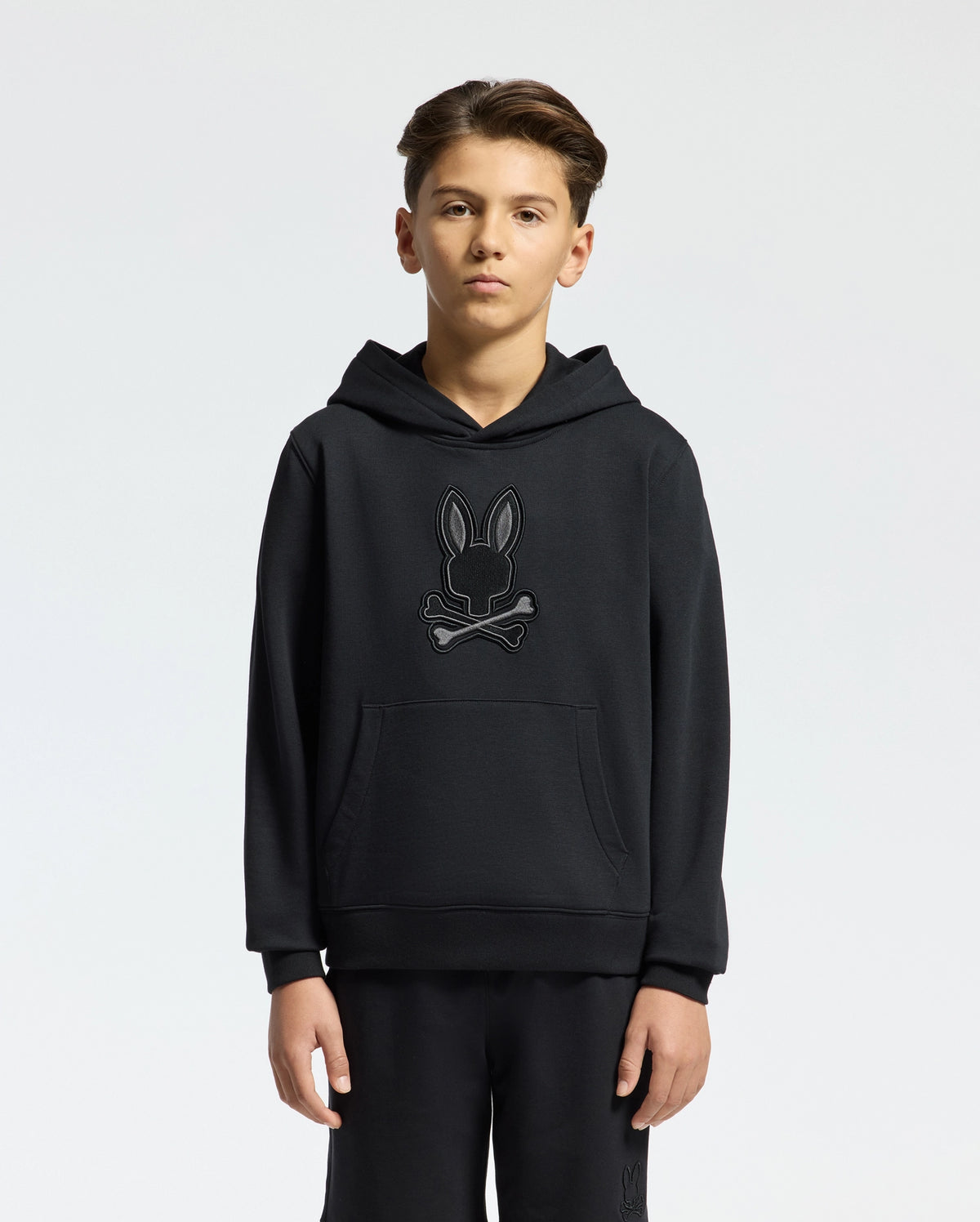A young person stands in a black Psycho Bunny KIDS NEW CASTLE FRENCH TERRY HOODIE, style B0H517F200, featuring an embroidered bunny and crossbones design on the front against a plain white background.