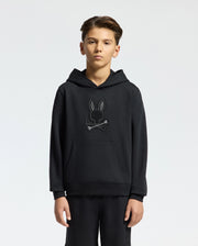A young person stands in a black Psycho Bunny KIDS NEW CASTLE FRENCH TERRY HOODIE, style B0H517F200, featuring an embroidered bunny and crossbones design on the front against a plain white background.