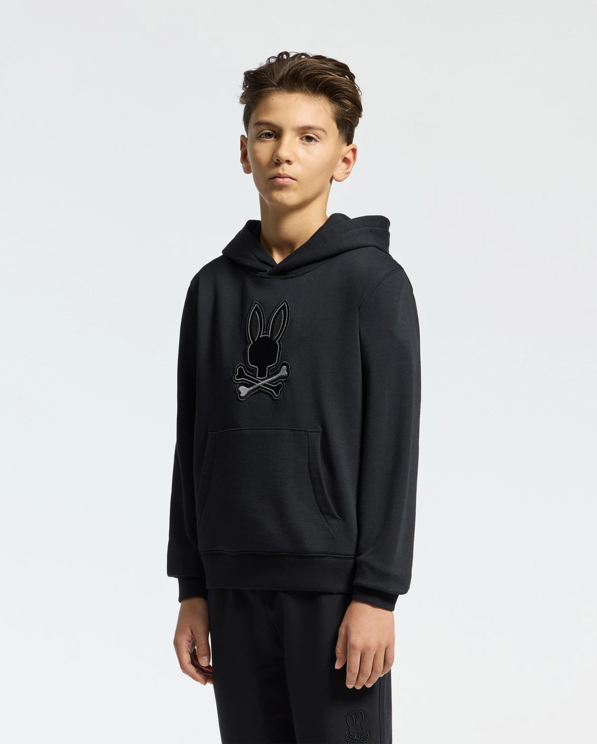 A young individual wears the KIDS NEW CASTLE FRENCH TERRY HOODIE - B0H517F200 by Psycho Bunny, adorned with an embroidered Bunny skull logo on the front against a plain white background.