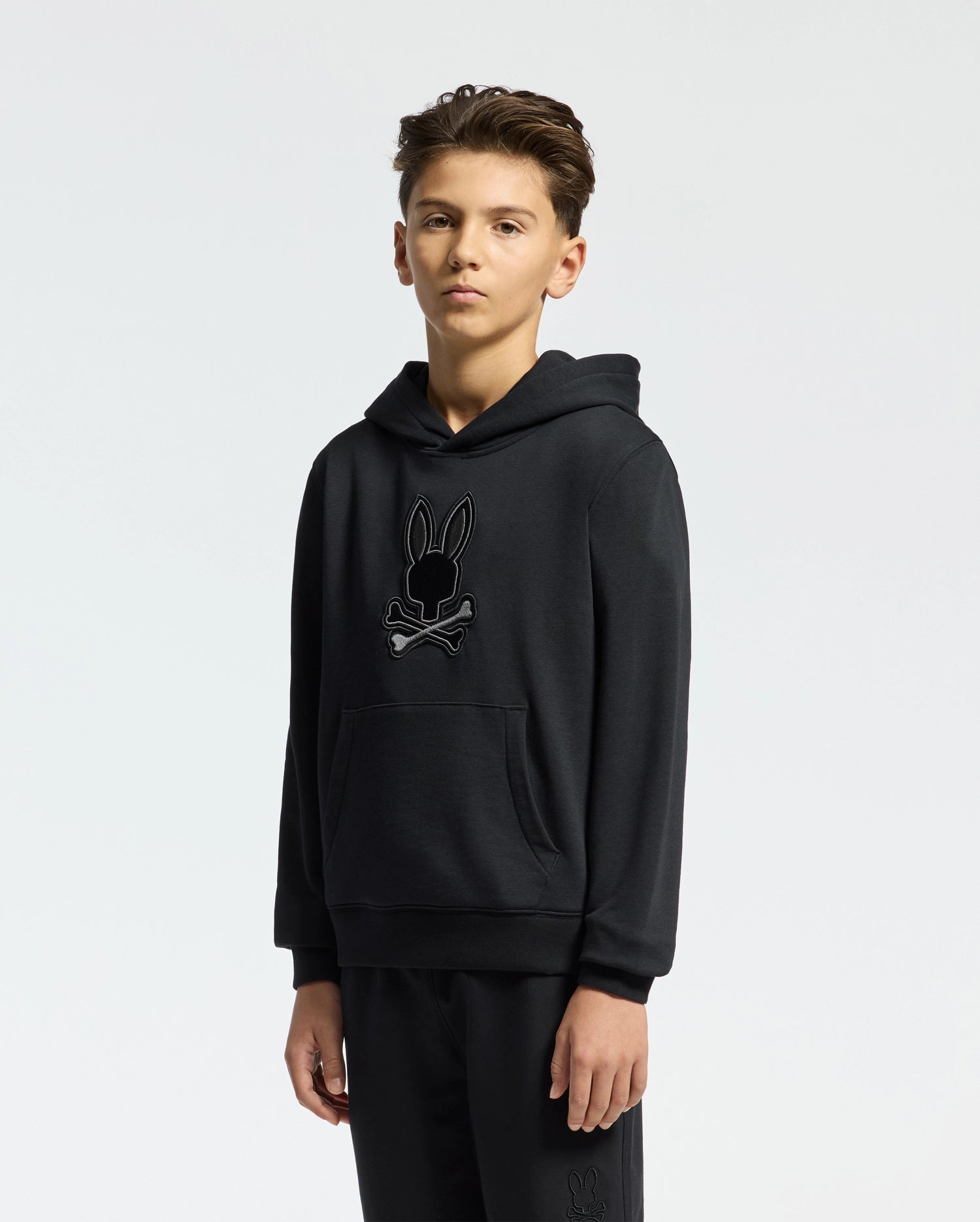A young individual wears the KIDS NEW CASTLE FRENCH TERRY HOODIE - B0H517F200 by Psycho Bunny, adorned with an embroidered Bunny skull logo on the front against a plain white background.
