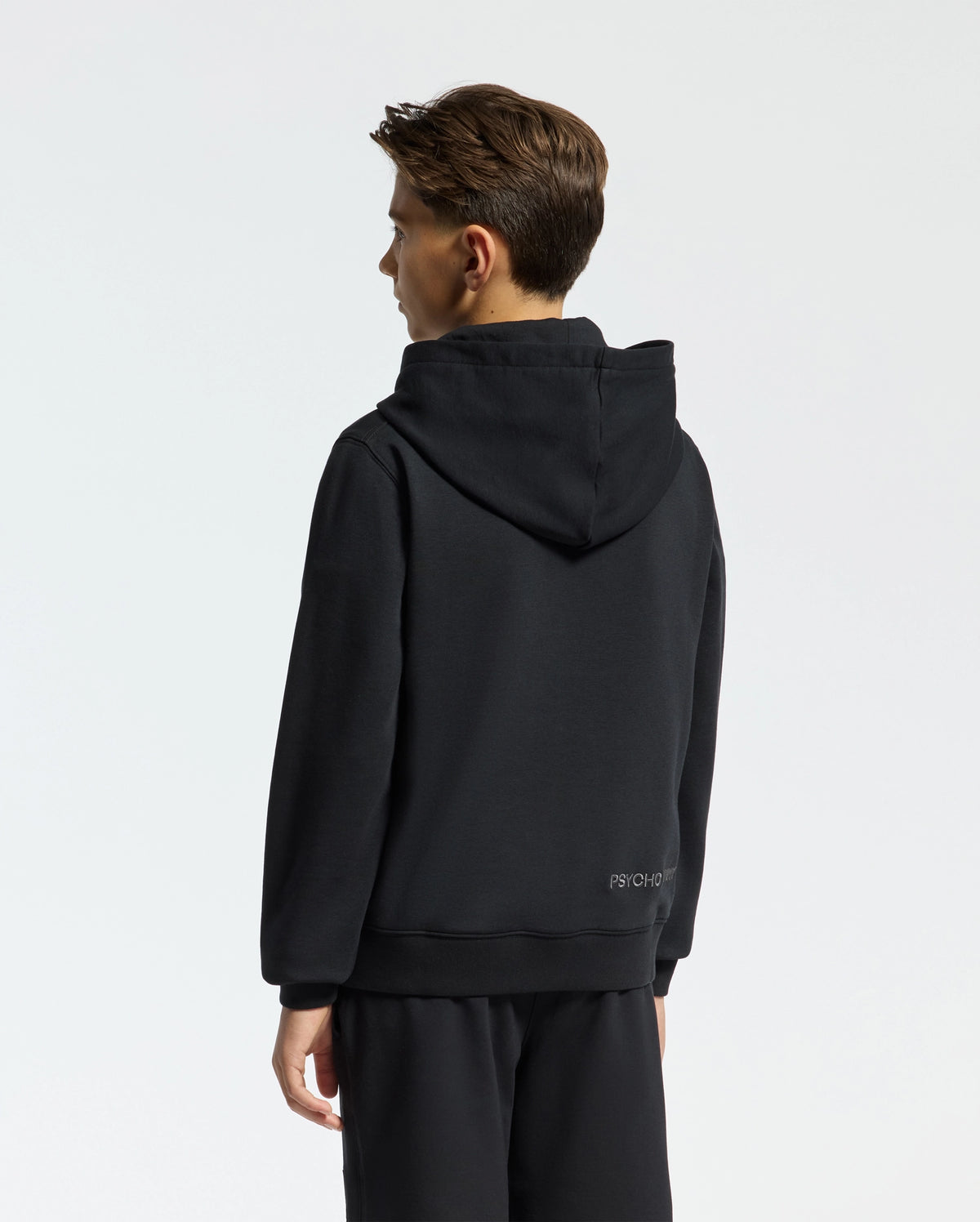 A young person in a Psycho Bunny black Kids New Castle French Terry Hoodie (B0H517F200) stands facing away, contrasting subtly against the plain, light-colored background.
