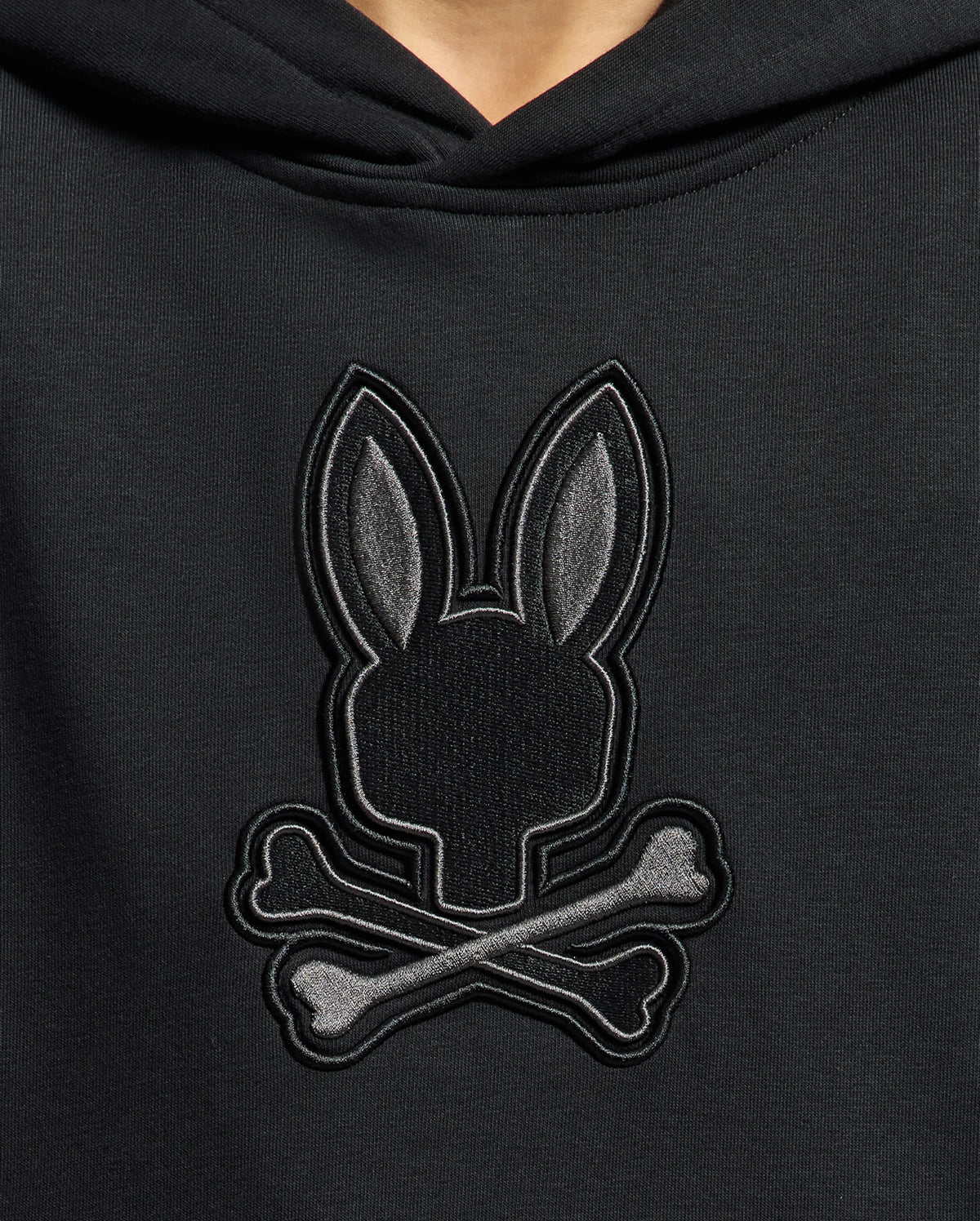Close-up of a Psycho Bunny KIDS NEW CASTLE FRENCH TERRY HOODIE featuring an embroidered emblem with a rabbit head and long ears over crossed bones, reminiscent of the iconic skull and crossbones motif.