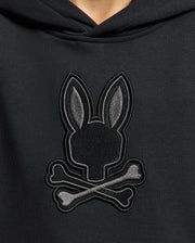 Close-up of a Psycho Bunny KIDS NEW CASTLE FRENCH TERRY HOODIE featuring an embroidered emblem with a rabbit head and long ears over crossed bones, reminiscent of the iconic skull and crossbones motif.