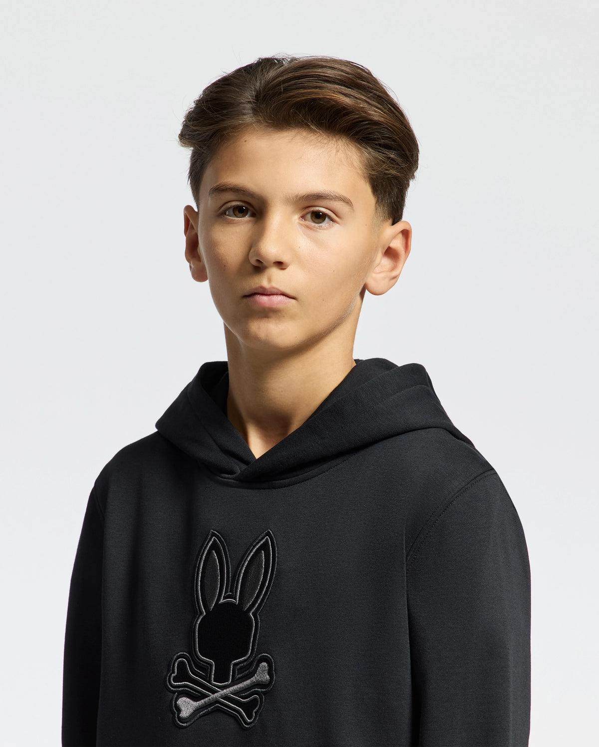 A young person with styled, short brown hair wears a Psycho Bunny KIDS NEW CASTLE FRENCH TERRY HOODIE in black, featuring an embroidered Bunny logo with a rabbit's head above crossbones, against a plain white background.
