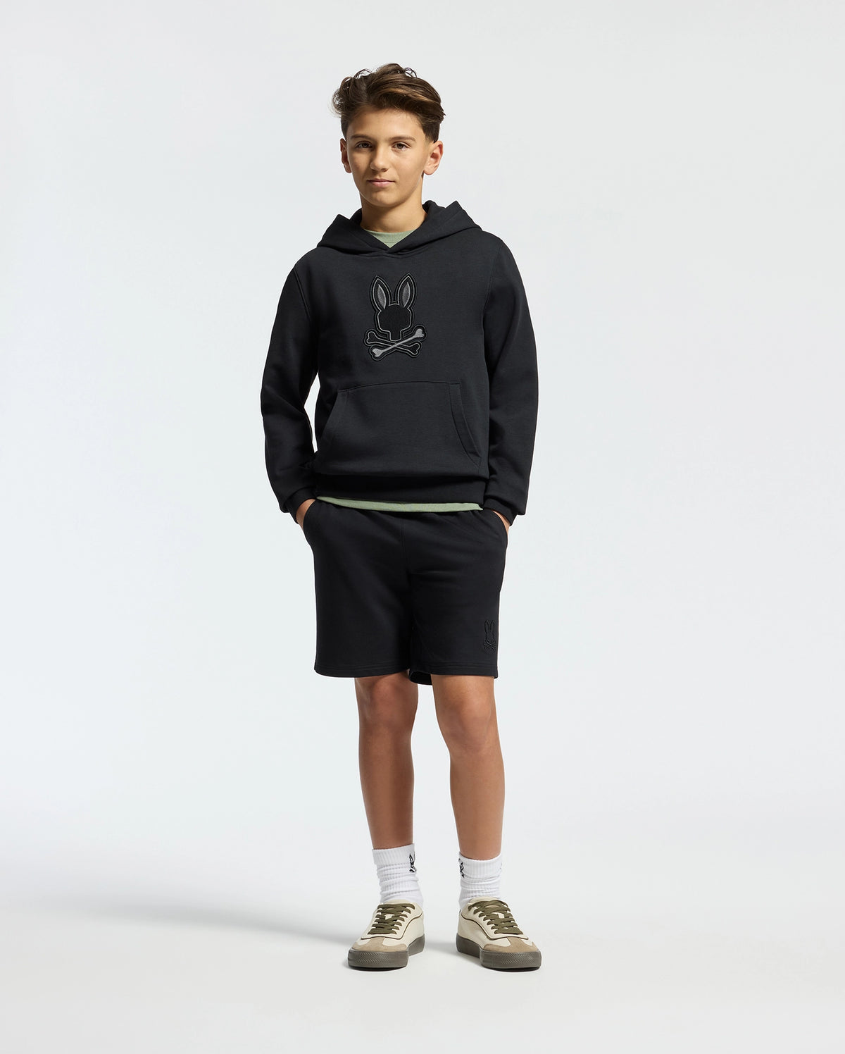 A teenage boy wears a Psycho Bunny KIDS NEW CASTLE FRENCH TERRY HOODIE, showcasing its embroidered Bunny logo. He pairs it with black shorts, white socks, and white sneakers featuring olive accents while standing casually with hands in pockets against a plain white background.