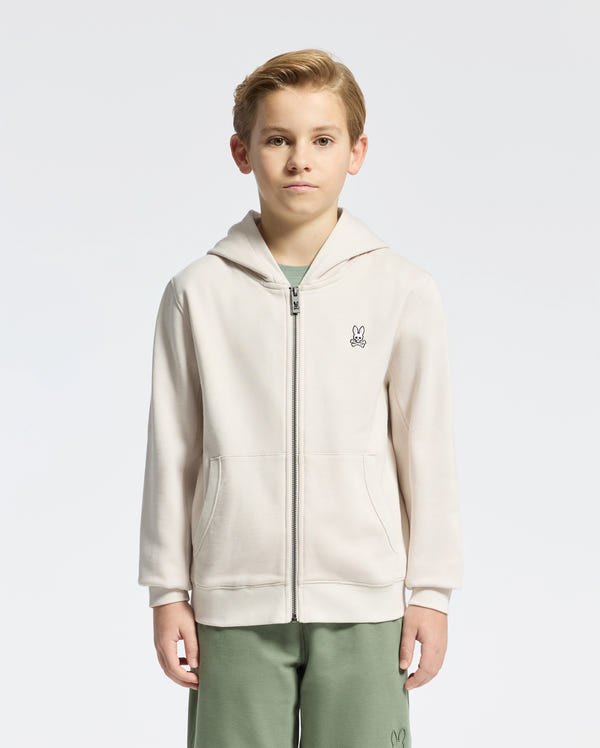 A young boy stands against a plain background wearing the KIDS LEVY ESSENTIAL ZIP FLEECE HOODIE by Psycho Bunny. Made from soft French terry cotton, it features a bunny logo on the chest. He pairs it with green pants, hands in pockets, looking directly at the camera.