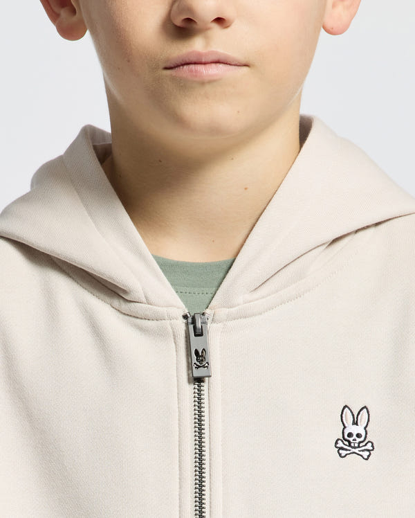 A child wears a partially unzipped Psycho Bunny Kids Levy Essential Zip Fleece Hoodie in light beige, revealing a green shirt underneath. The hoodie features a zipper and an embroidered bunny logo on the chest. The image captures the upper torso and part of the face.
