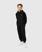 A young boy stands smiling, wearing a regular fit black Psycho Bunny Kids Levy Fleece Hoodie and matching sweatpants with a subtle logo. He has brown hair, white sneakers, and the outfit is crafted from 100% cotton. The background is plain and light-colored.