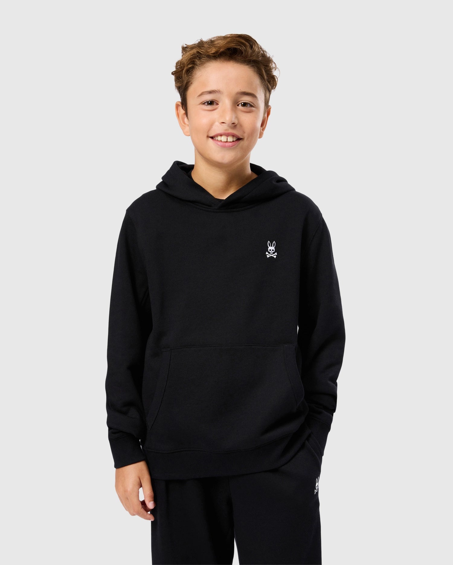 The boy with short brown hair is smiling while wearing a black, regular fit Kids Levy Fleece Hoodie by Psycho Bunny, featuring a small white logo on the chest. He stands against a plain light gray background.