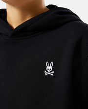 A person is wearing the KIDS LEVY FLEECE HOODIE - B0H859D200 by Psycho Bunny, a regular fit black hoodie crafted from 100% cotton. It features a small embroidered white logo of a bunny with crossed bones underneath on the chest, adding a playful twist to the traditional skull and crossbones design.