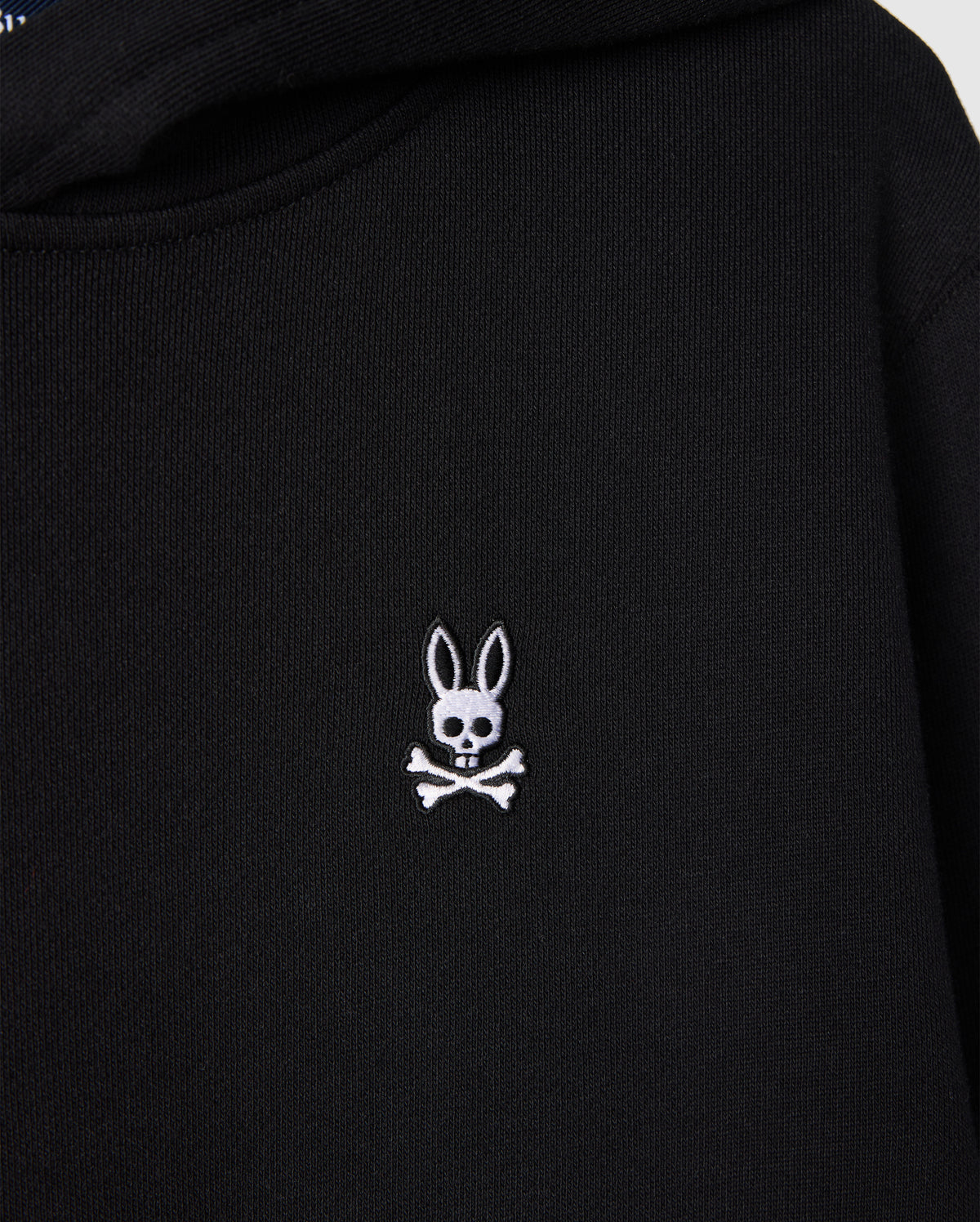 The Psycho Bunny KIDS LEVY FLEECE HOODIE in black displays a distinctive small white embroidered rabbit's head with long ears, artistically positioned over a skull and crossbones. Crafted from 100% cotton, this hoodie offers a regular fit that combines uniqueness with comfort.