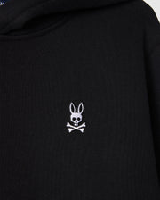The Psycho Bunny KIDS LEVY FLEECE HOODIE in black displays a distinctive small white embroidered rabbit's head with long ears, artistically positioned over a skull and crossbones. Crafted from 100% cotton, this hoodie offers a regular fit that combines uniqueness with comfort.