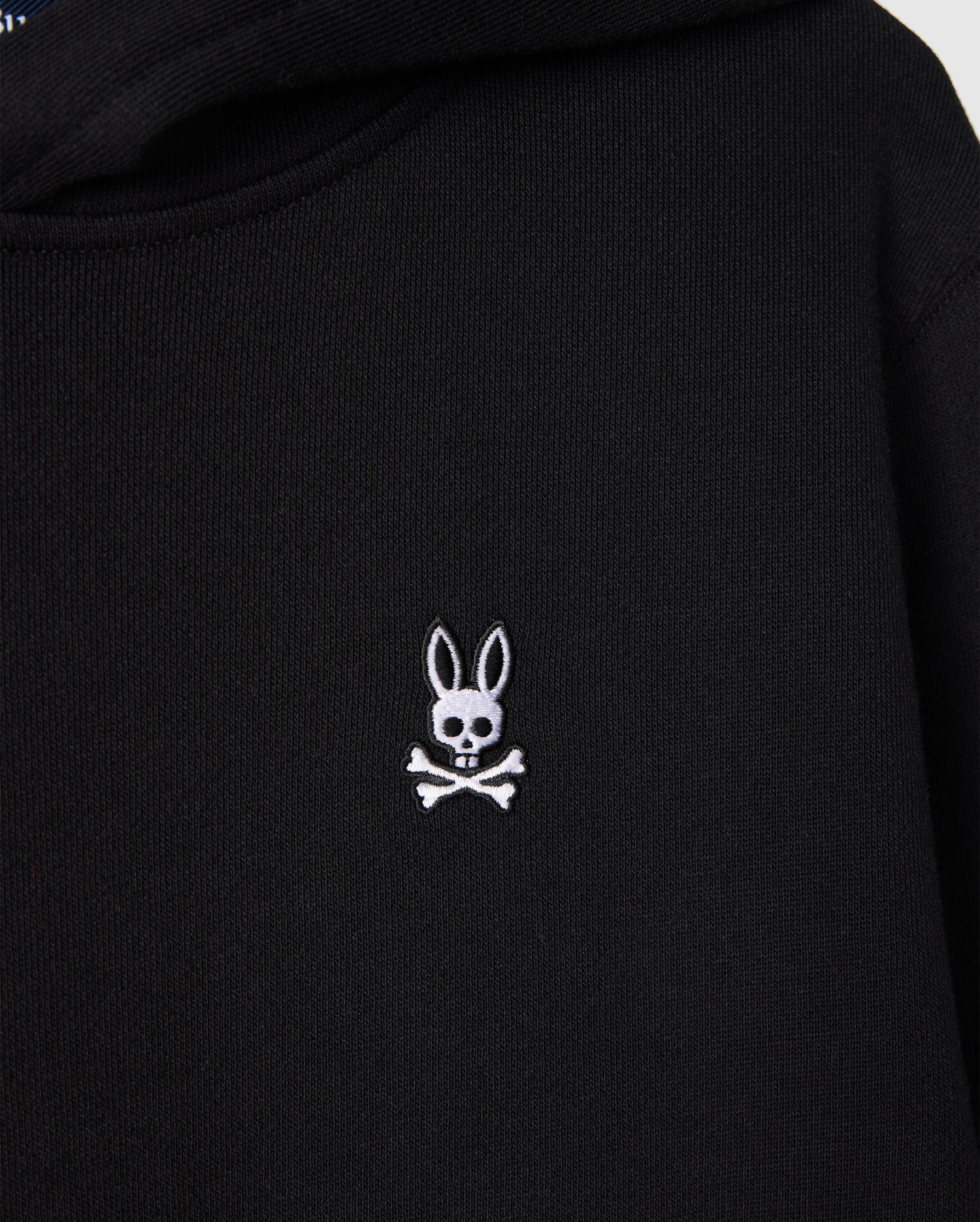 The Psycho Bunny KIDS LEVY FLEECE HOODIE in black displays a distinctive small white embroidered rabbit's head with long ears, artistically positioned over a skull and crossbones. Crafted from 100% cotton, this hoodie offers a regular fit that combines uniqueness with comfort.